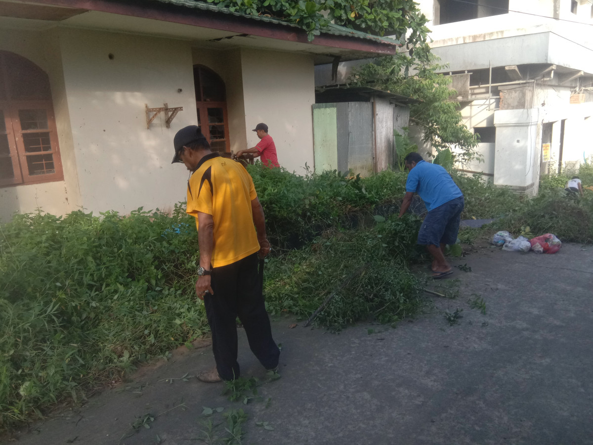 Menebas rumput dan tanaman liar di sepanjang jalan poros dan membersihkan parit yg tersumbat sampah